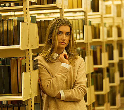 Elena pensively leans against a bookshelf in the academic section of a public library.
