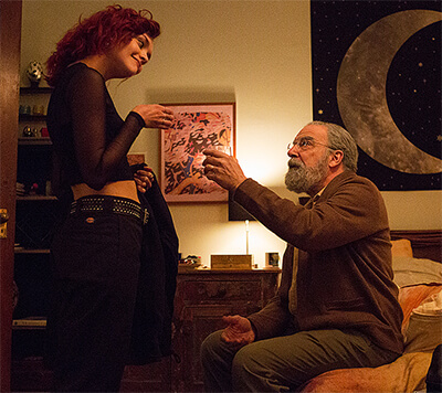 Dylan, standing, raises her glass to Irwin; sitting on a bed and raising his glass in return.