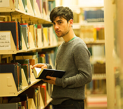 Rodrigo stands, holding a book but looking elsewhere, by a bookshelf in the academic section of a public library.