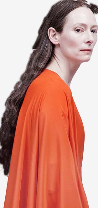 Madame Blanc stands, slightly hunched, in orange robes looking at the camera with a superior experession.