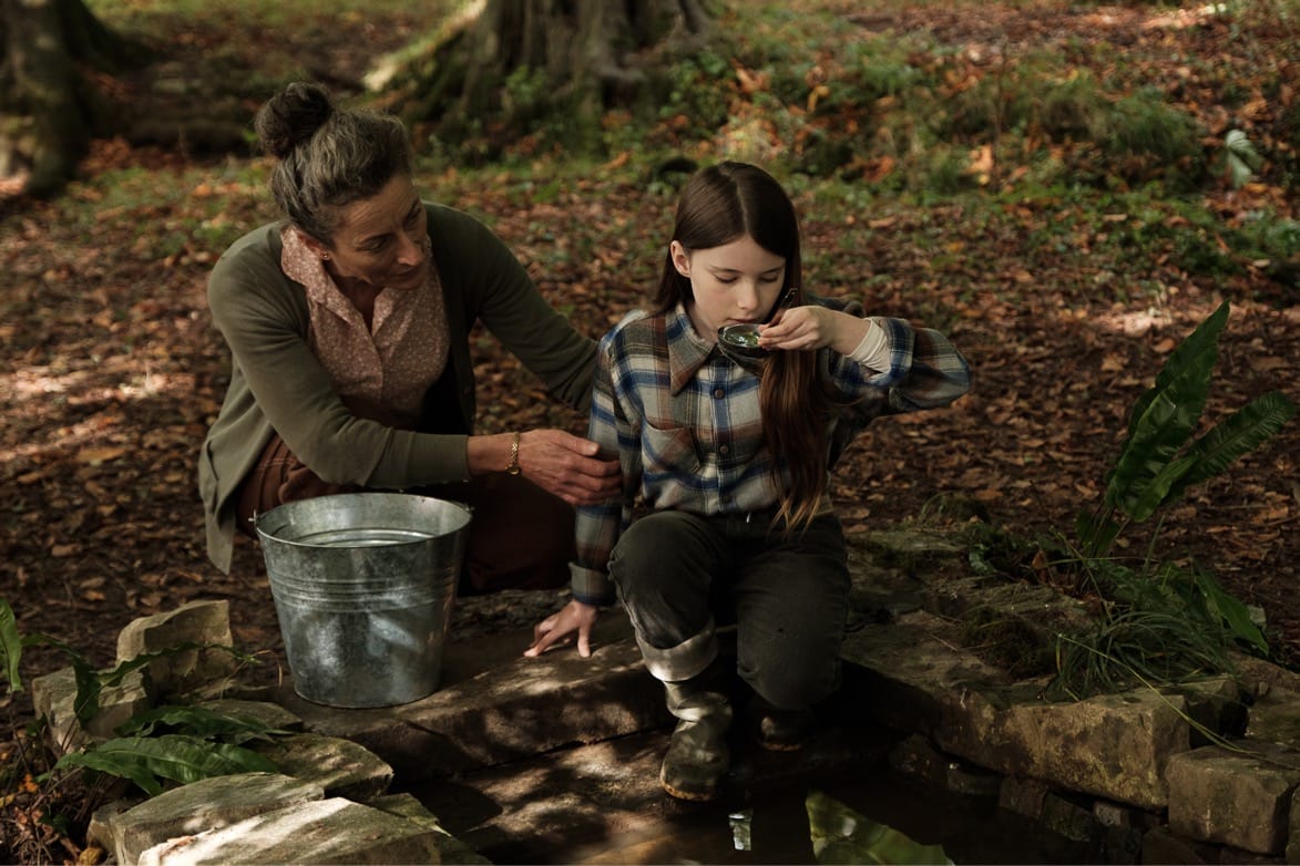 In the woods, Carrie Crowley holds the child Catherine Clinch's arm as she drinks a ladle of water from a bucket.