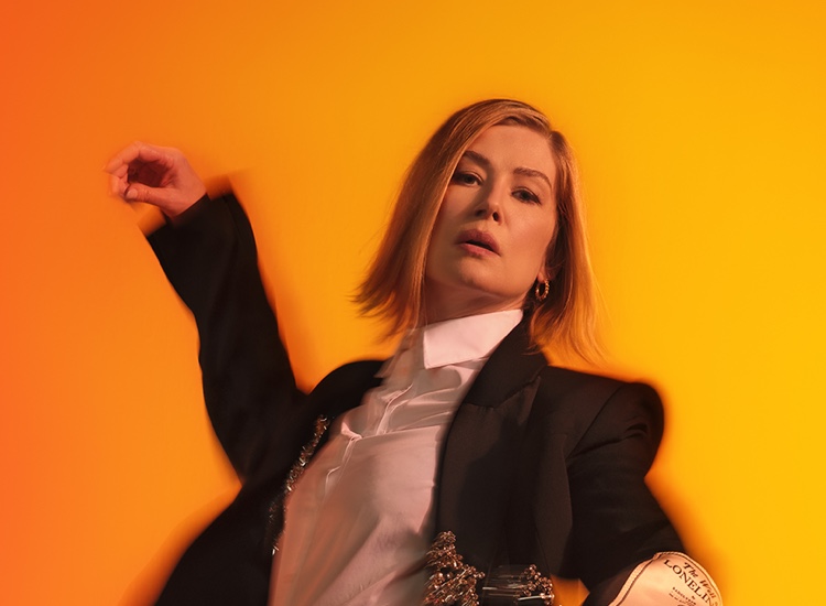 Rosamund Pike stands in a suit with her arms in the air, in front of an orange background.