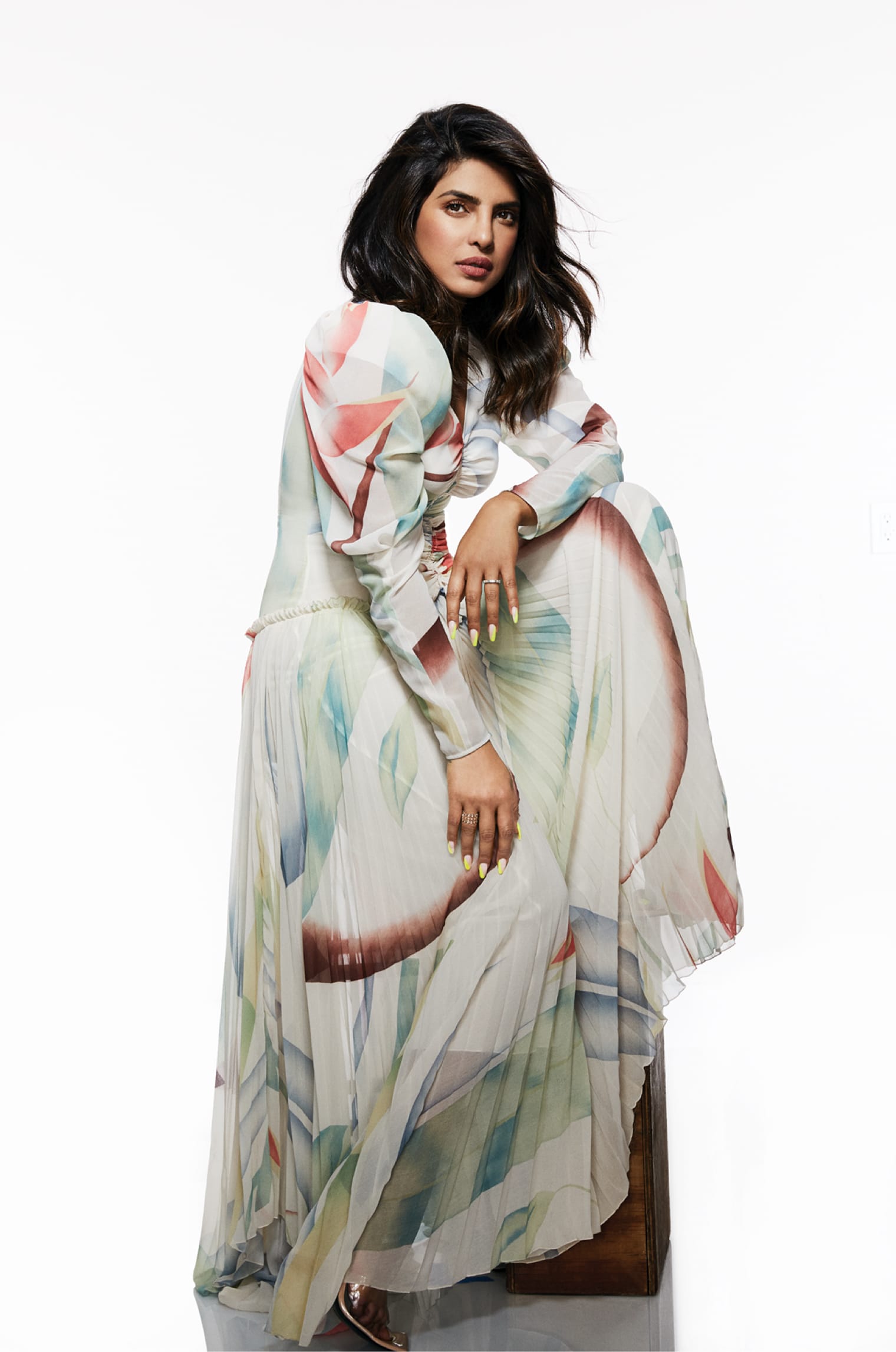 Priyanka Chopra Jonas poses perched on a seat. The skirt of her colorful dress cascades like a waterfall. Her manicure is impeccable.